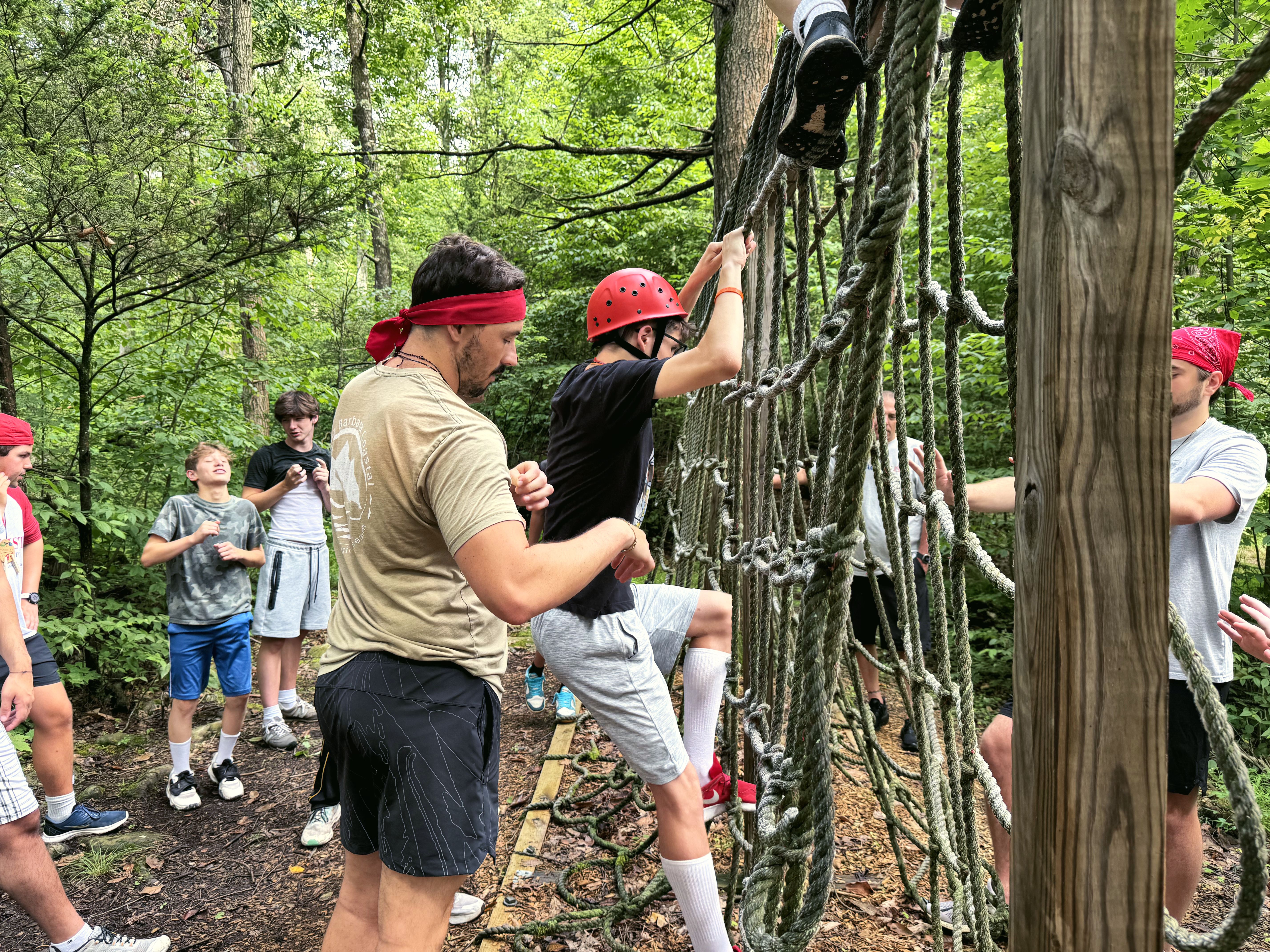 A young leader spots a camper climbing a rope net on the ropes course while others watch