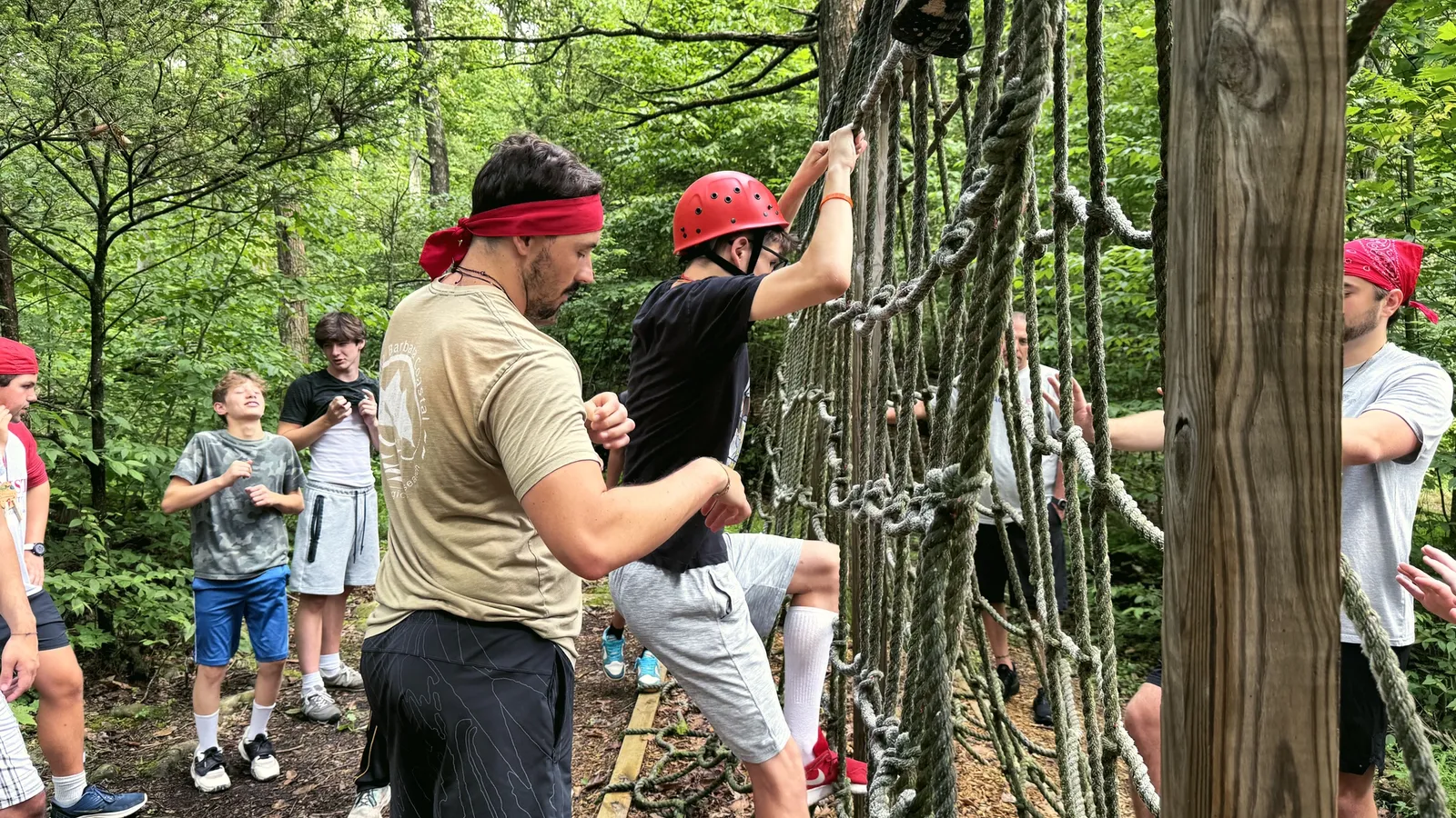 A young leader spots a camper climbing a rope net on the ropes course while others watch