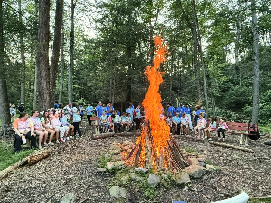 Campers and counselors sit on logs around a big campfire in a forest clearing.