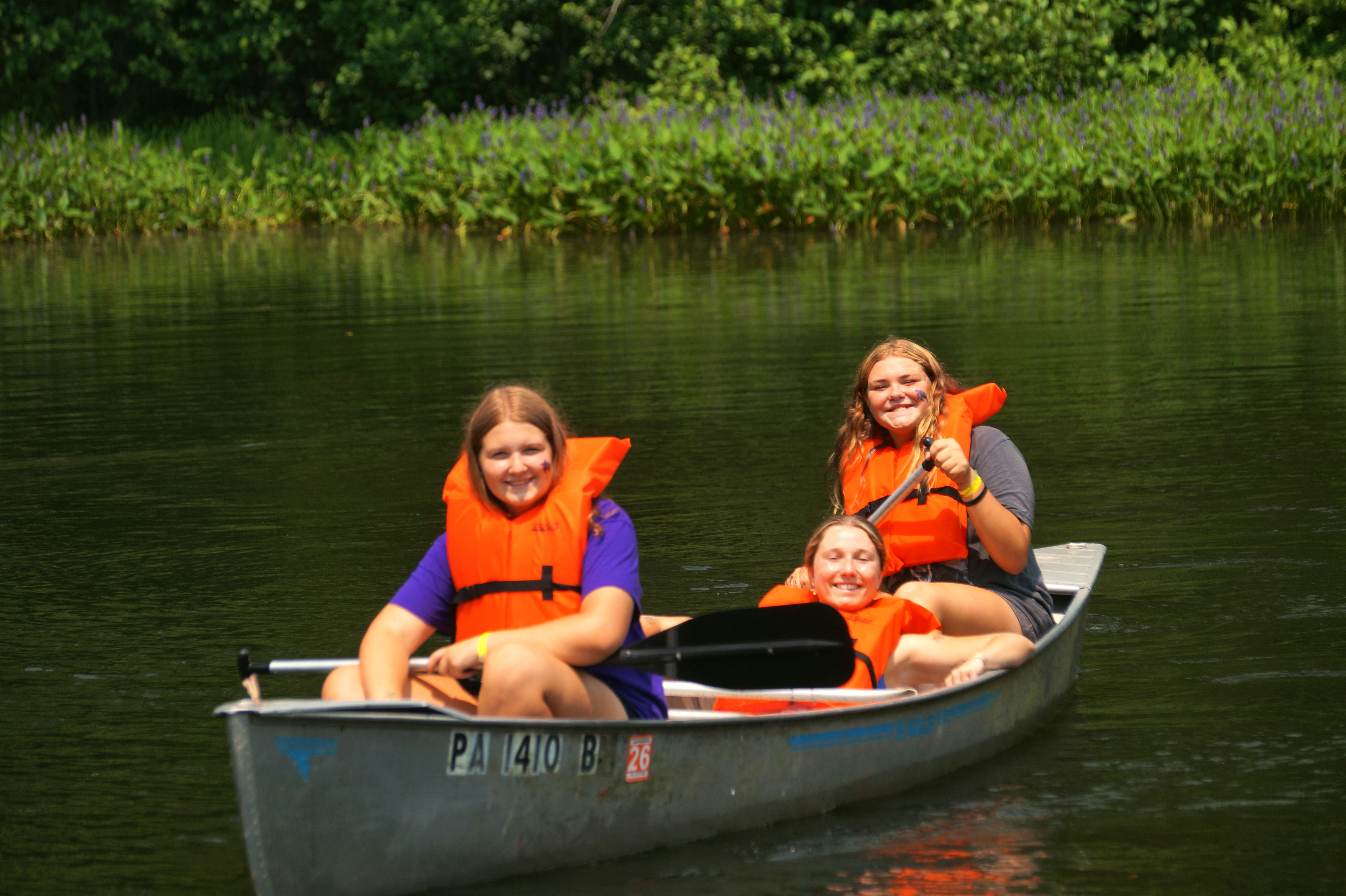 Three campers in orange life vests smile as they paddle a canoe down a calm river.