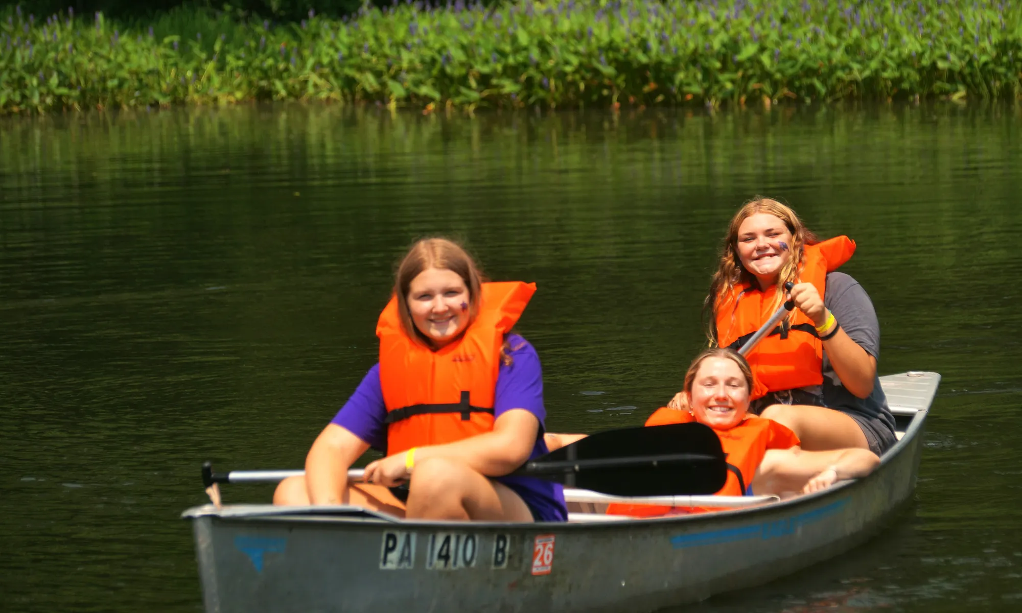 Three campers in orange life vests smile as they paddle a canoe down a calm river.