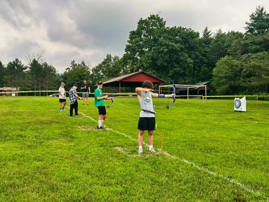Teens practice archery on a grassy field, aiming at targets