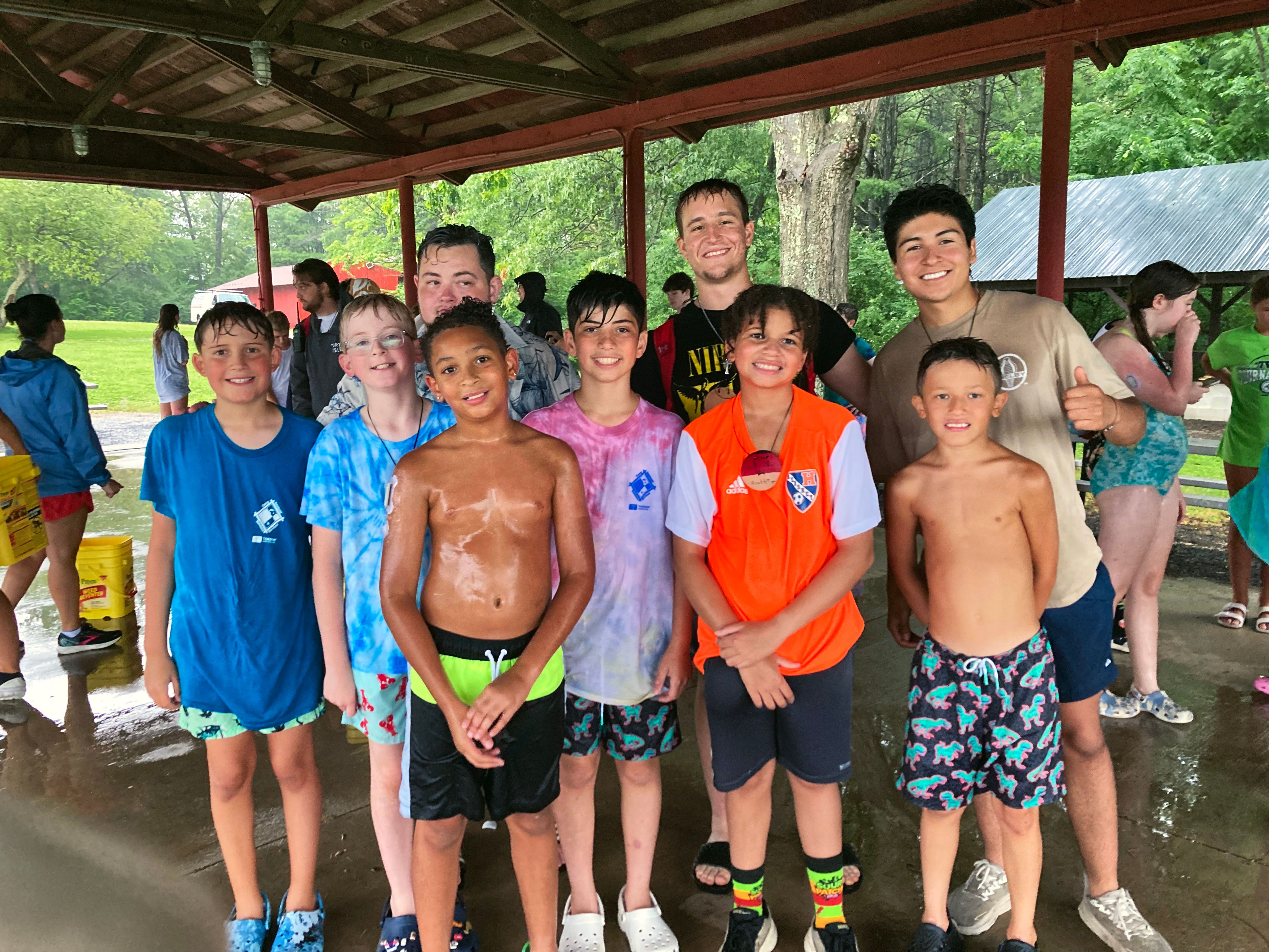 Two male counselors stand with a group of smiling young campers under a covered pavilion