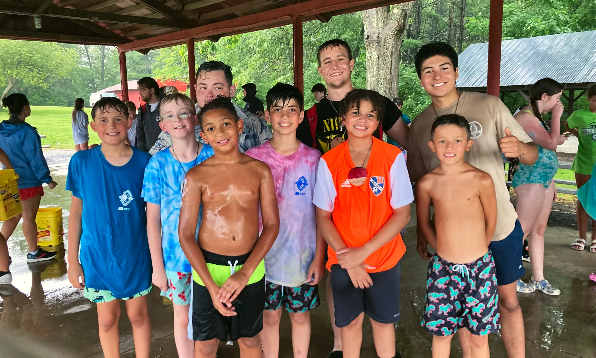 Two male counselors stand with a group of smiling young campers under a covered pavilion