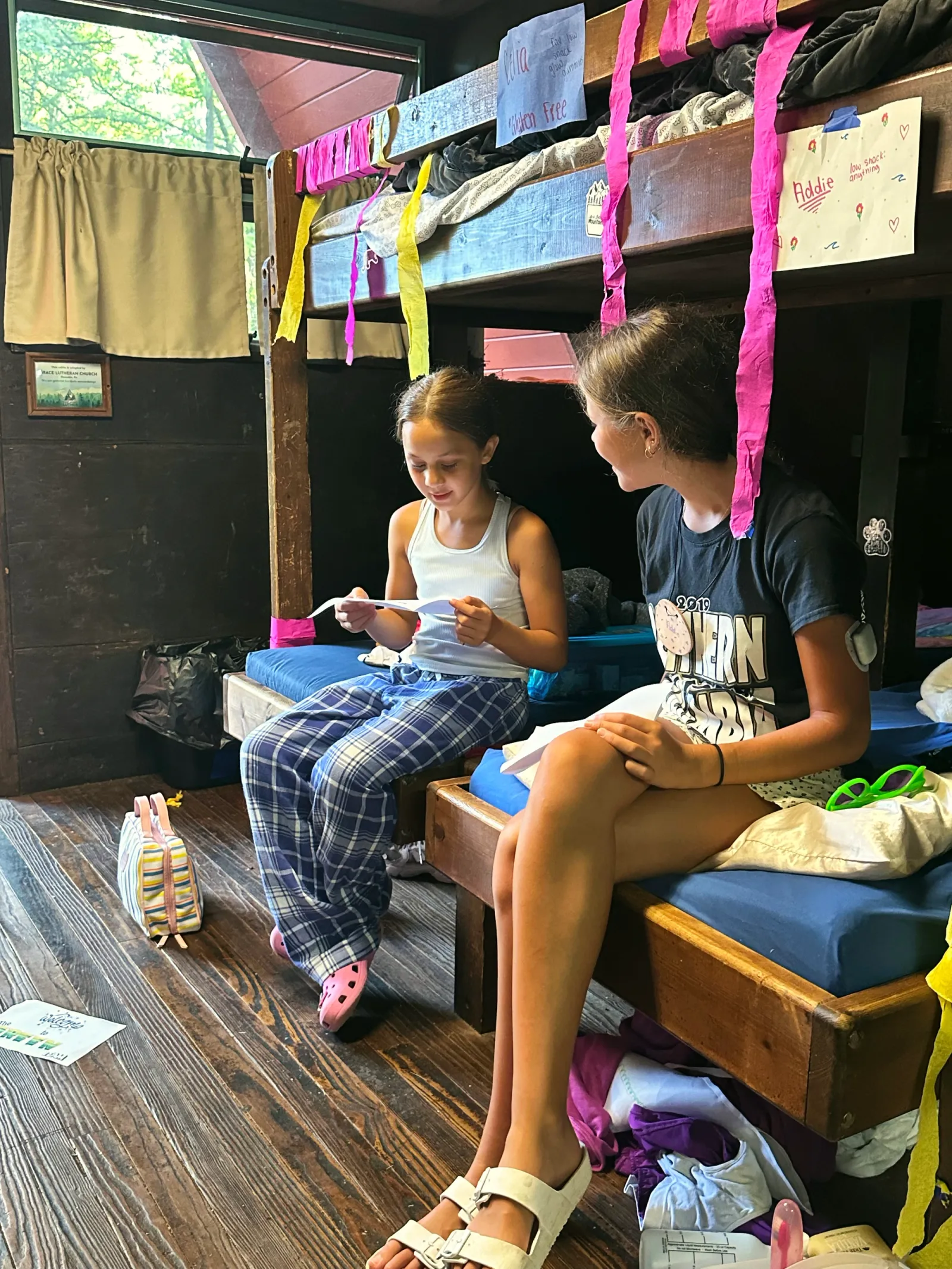 Two girls sit on a bunk bed in a rustic cabin decorated with ribbons
