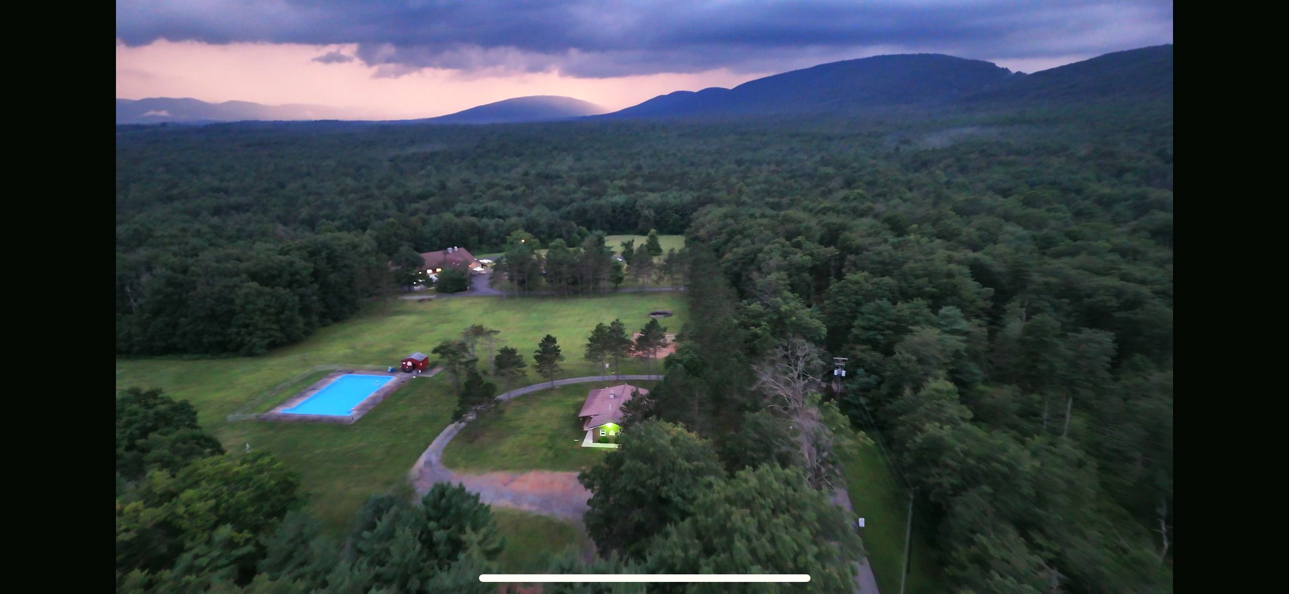 Aerial view of the camp at dusk, with a swimming pool and a few buildings in a clearing in the forest