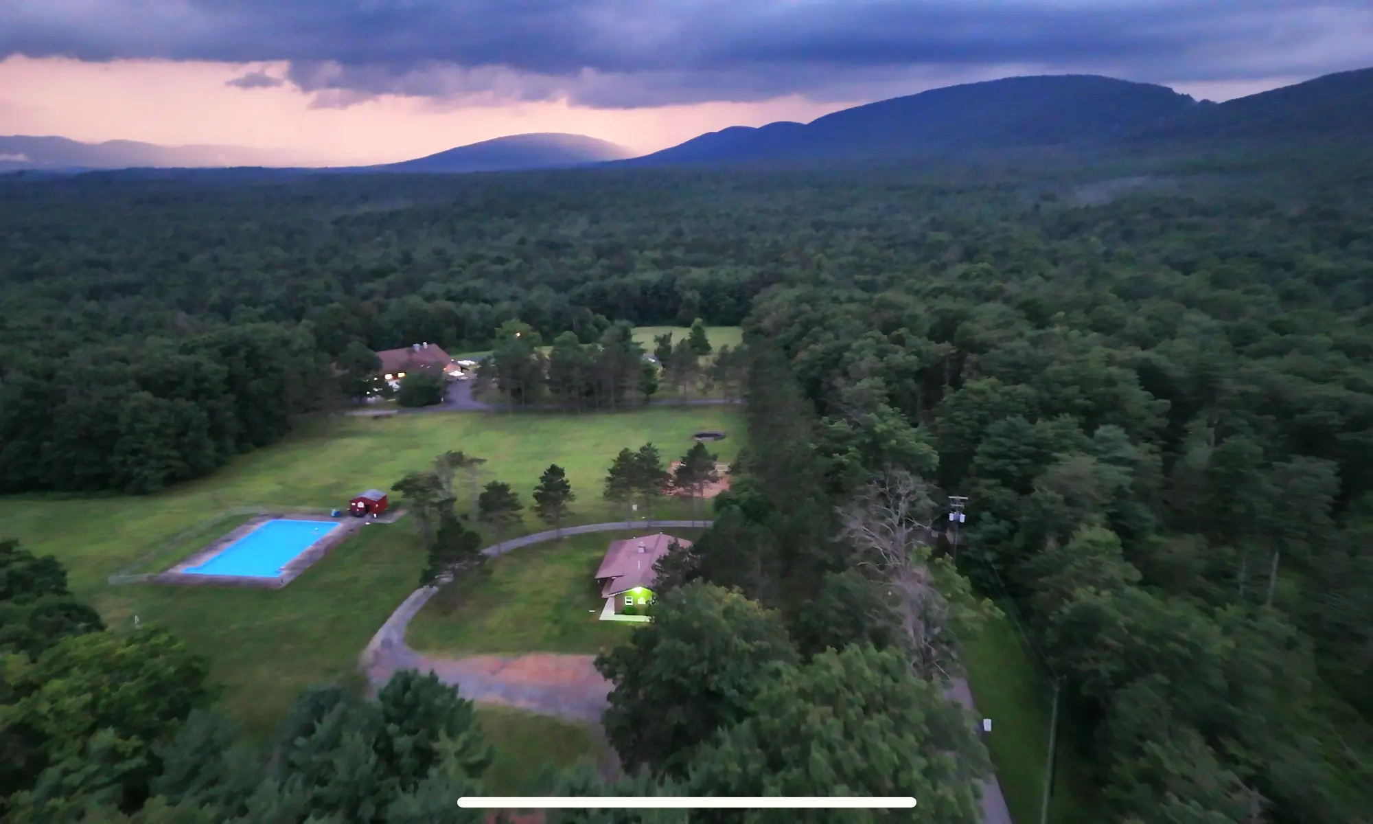 Aerial view of the camp at dusk, with a swimming pool and a few buildings in a clearing in the forest