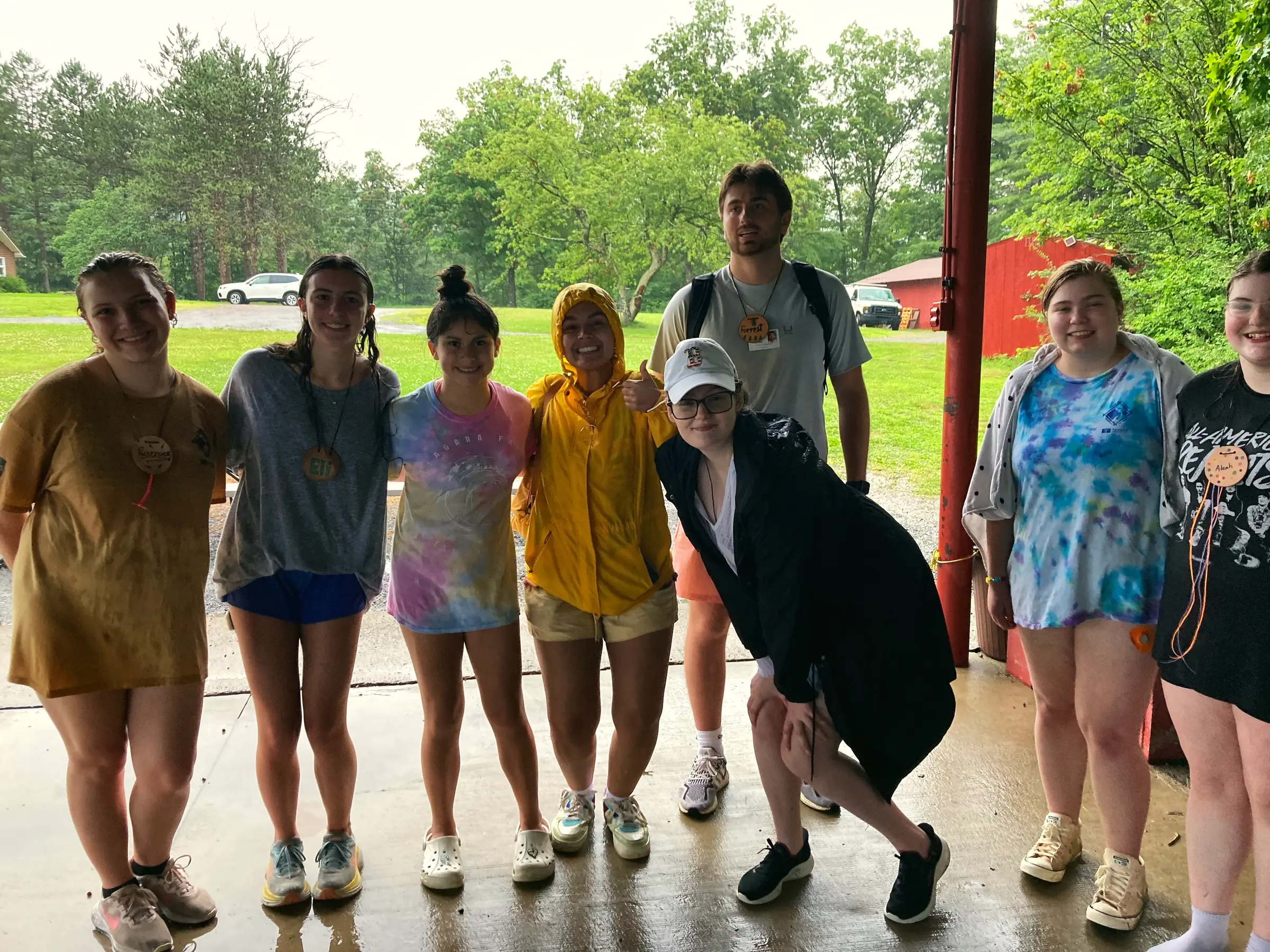 Eight young staff members, some in tie-dye shirts and one in a yellow raincoat, pose together under a covered porch on a rainy day