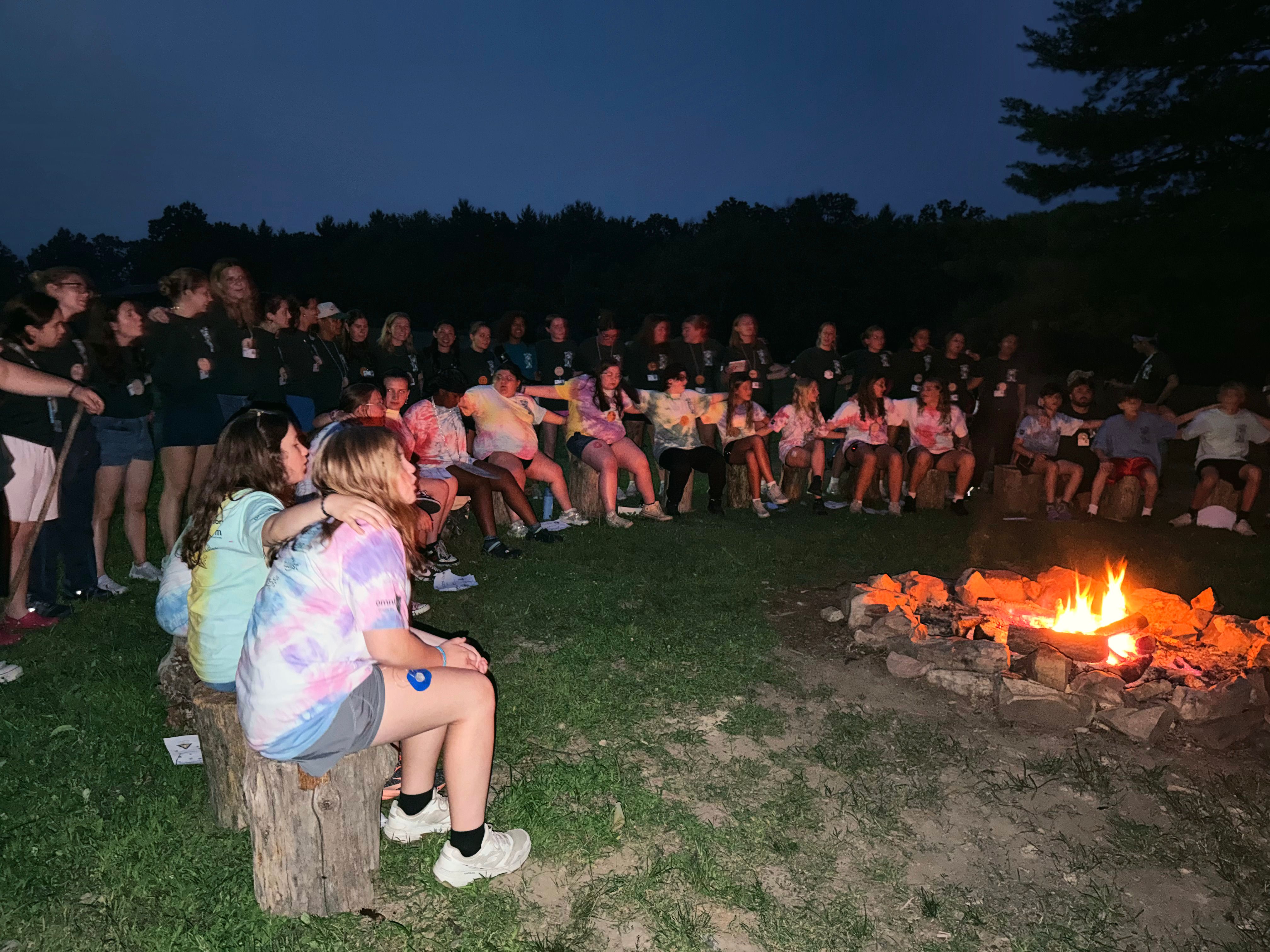 Campers sit in a circle around a campfire at dusk