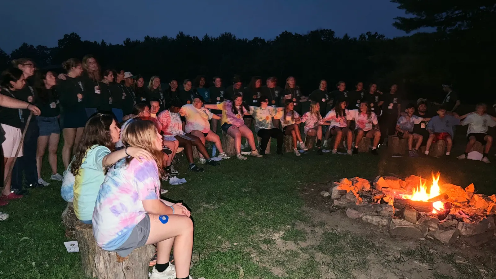 Campers sit in a circle around a campfire at dusk