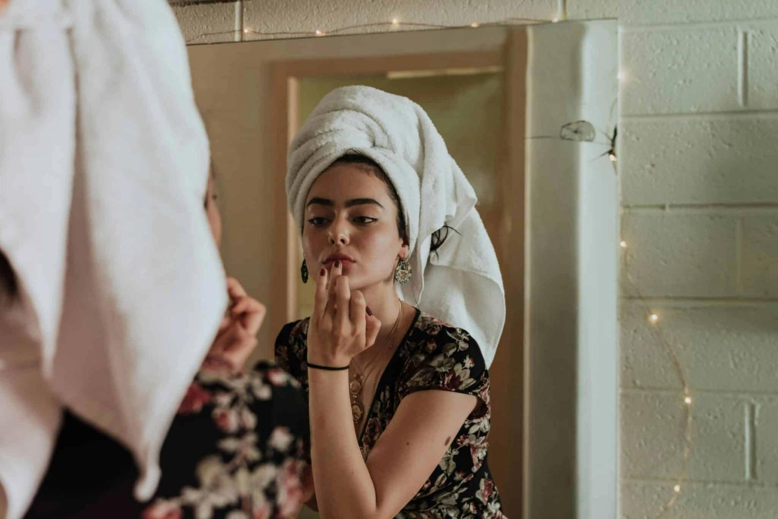 Woman with hair wrapped in a towel applying makeup in a mirror