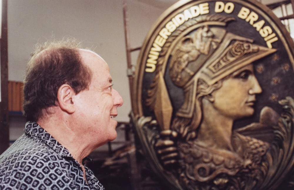 Fotografia de Joaquim de Lemos e Souza de perfil, sorridente, olhando para um grande medalhão dourado da Universidade do Brasil. O medalhão traz o brasão da UFRJ com a figura da deusa Minerva em relevo, com capacete e detalhes ornamentais.