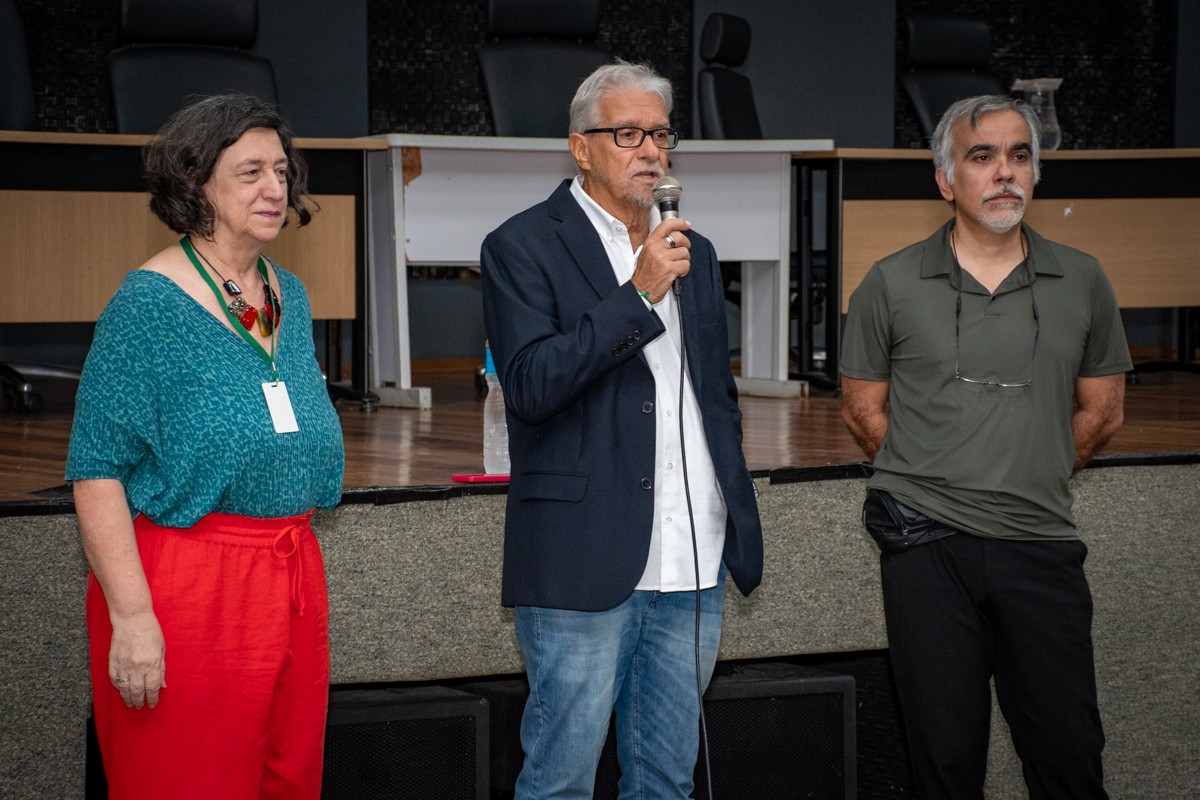 A imagem mostra três pessoas de pé em um auditório, em frente a uma mesa longa com cadeiras pretas, durante um evento acadêmico.
À esquerda está Lina Zingali, vice-decana do Centro de Ciências da Saúde (CCS), com cabelos curtos e ondulados, grisalhos, vestindo uma blusa verde-azulada estampada, calça vermelha e um colar grande vermelho. Ela usa um crachá pendurado no pescoço. No centro está Luis Eurico Nasciutti, decano do CCS, de cabelos brancos e óculos, vestindo um blazer azul-marinho sobre uma camisa branca e calça jeans azul. Ele segura um microfone e parece estar falando ao público. À direita está Marcus Oliveira, professor do Instituto de Bioquímica Médica Leopoldo de Meis (IBqM/UFRJ), de cabelos grisalhos e barba curta, vestindo uma camisa polo verde-escura e calça preta, com óculos pendurados na gola. Ele está com as mãos para trás, em posição relaxada.