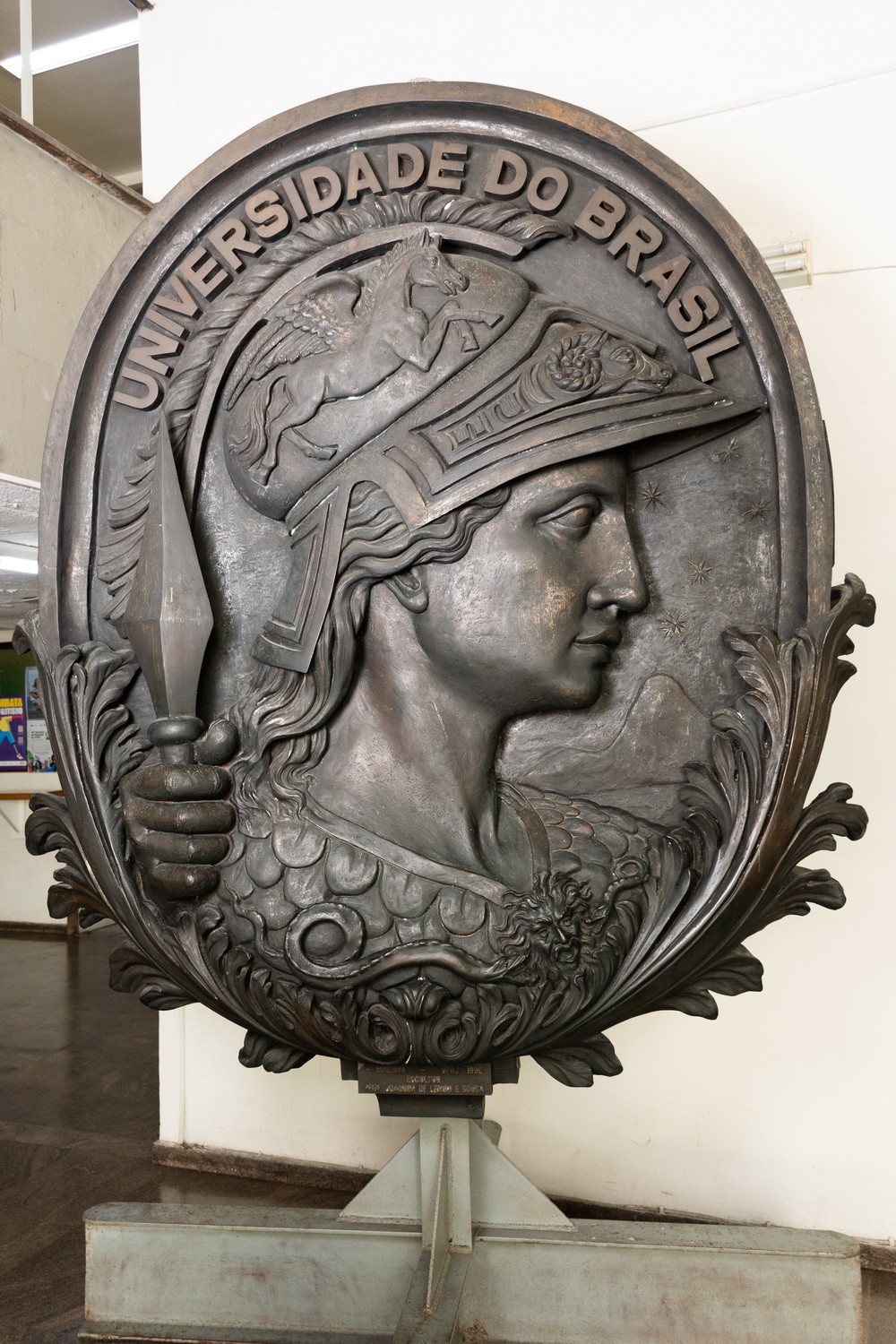 Medalhão do brasão localizado no bloco K do CCS que traz a Minerva em destaque, em tom dourado, com a inscrição Universidade do Brasil. A deusa está em perfil, usando capacete com adornos e segurando uma lança.