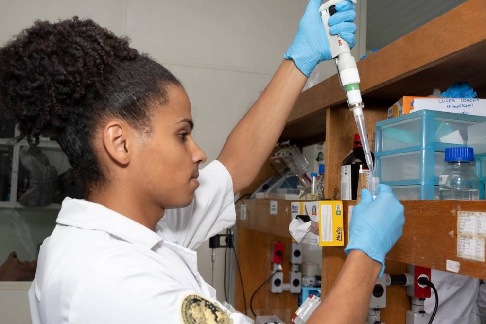 Um jovem negro usando jaleco branco e luvas azuis manuseia uma micropipeta em um laboratório. Ele transfere líquido para um tubo de ensaio posicionado em uma bancada de madeira com caixas organizadoras, reagentes e materiais de pesquisa. O ambiente é bem iluminado e típico de um laboratório de ciências biológicas.