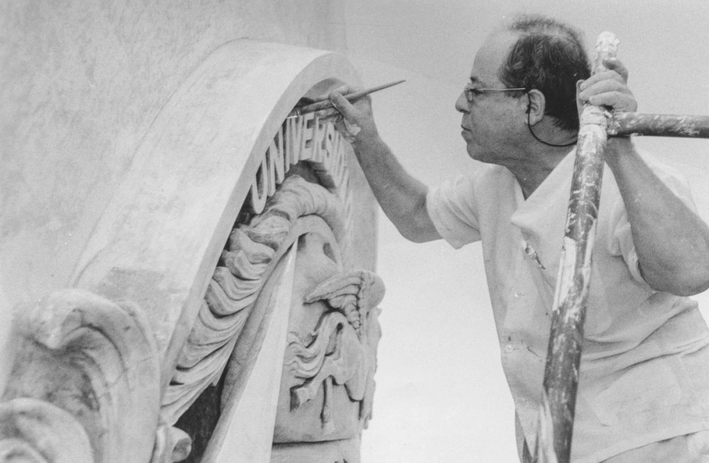 Fotografia de Joaquim de Lemos trabalhando na escultura do brasão com a deusa Minerva. Ele aparece em processo de confecção da peça, utilizando ferramentas de escultura.