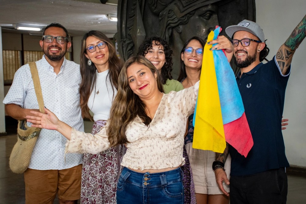 Sete pessoas estão posando felizes e sorrindo, voltadas diretamente para a câmera. Elas estão próximas umas das outras, demonstrando amizade ou espírito de grupo. No centro, uma mulher segura uma bandeira colorida com três faixas: amarela, azul e vermelha (correspondente à bandeira da Colômbia).
