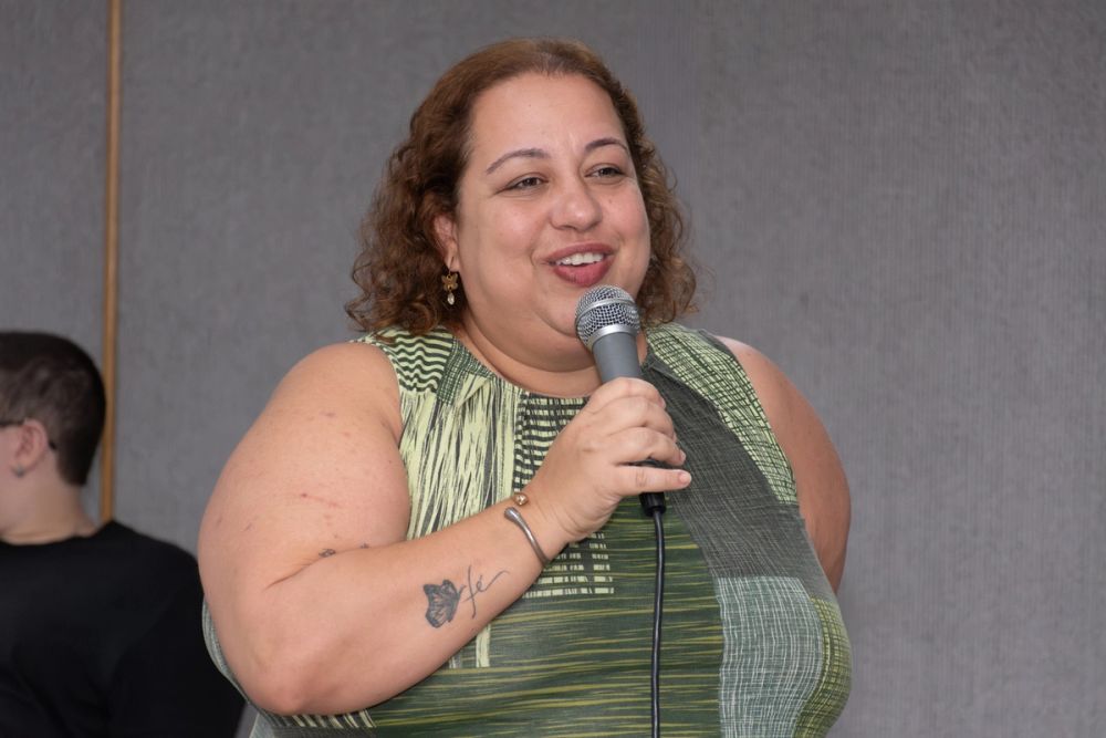 Na imagem há duas pessoas em ambiente interno. Em primeiro plano, ao centro e ocupando a maior parte do enquadramento, aparece a professora Avany Fernandes, uma mulher adulta, em faixa etária aproximada entre 45 e 55 anos, de pele clara e cabelos castanhos curtos e cacheados, na altura dos ombros. O rosto está voltado levemente para a direita da imagem, com olhar direcionado para o mesmo lado e boca entreaberta. A pessoa veste blusa sem mangas em tons de verde-claro, verde-oliva e verde-escuro, com estampas geométricas e linhas horizontais e verticais. Usa brincos pequenos dourados com pingente claro, pulseira metálica no punho direito e segura com a mão direita um microfone cinza com tela metálica prateada e cabo preto, posicionado próximo à boca. No antebraço direito há tatuagens visíveis em tinta escura: uma próxima ao cotovelo e outra mais central, com desenho de flor e traços finos. O braço esquerdo está voltado para trás do corpo e não aparece completamente. À esquerda da imagem, em segundo plano, aparece parcialmente outra pessoa, visível do ombro para cima, com cabelo bem curto escuro, pele clara e blusa preta, posicionada de perfil voltado para a esquerda. O fundo é formado por grandes painéis cinza-claro e cinza-médio com textura vertical discreta e uma linha de divisão em tom bege à esquerda. A iluminação é uniforme e clara, sem sombras intensas. O enquadramento é horizontal, do quadril para cima da pessoa em primeiro plano. Não há textos, logotipos ou outros elementos gráficos visíveis. Predominam na imagem os tons de cinza do fundo, verde da roupa e cinza metálico do microfone.