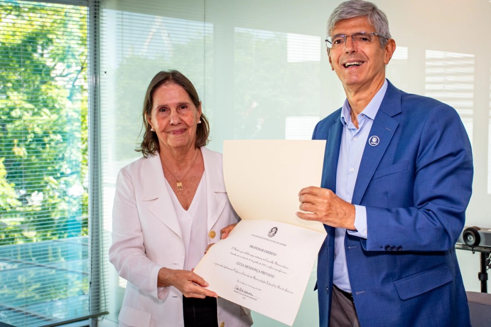 A professora Lúcia Mendonça Previato e o reitor da UFRJ, Roberto Medronho, posam lado a lado em uma sala iluminada por uma janela com persianas e vista para árvores. Medronho, de terno azul, segura e apresenta o diploma do título de Professora Emérita concedido à docente, enquanto Lúcia, vestindo blazer claro, apoia o documento e sorri para a câmera.