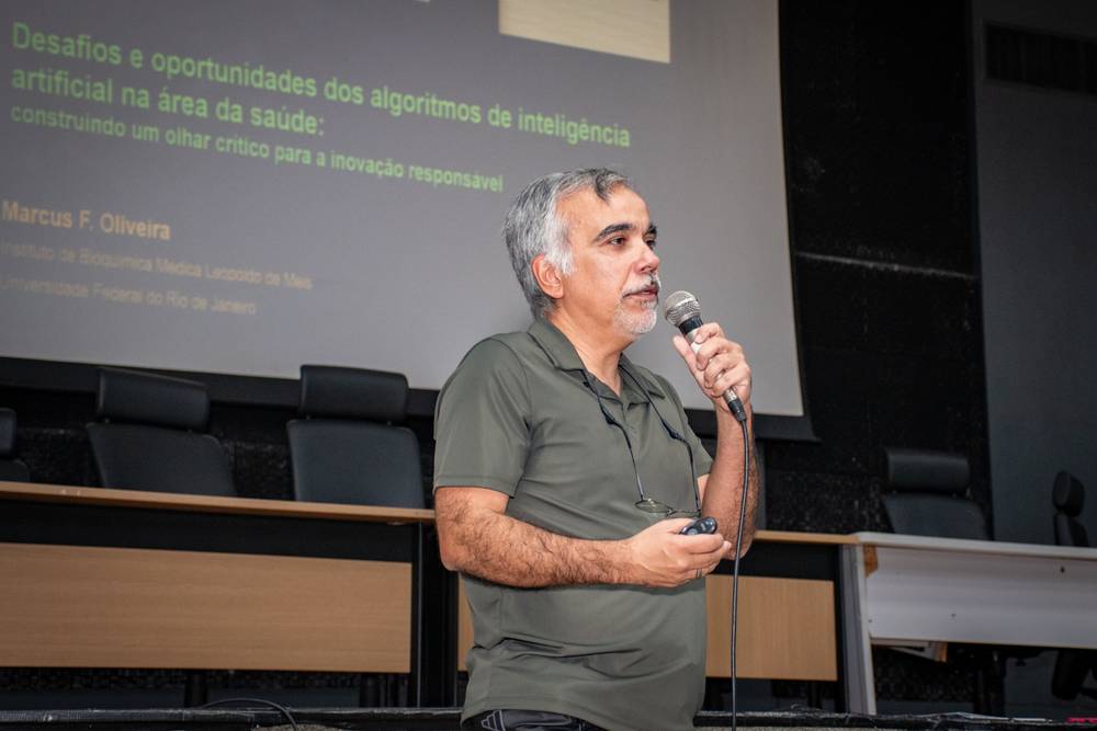 A imagem mostra três pessoas de pé em um auditório, em frente a uma mesa longa com cadeiras pretas.
À esquerda, está uma mulher de cabelos curtos e ondulados, grisalhos, vestindo uma blusa verde-azulada estampada, calça vermelha e um colar grande vermelho. Ela usa um crachá pendurado no pescoço.
No centro, está um homem de cabelos brancos e óculos, vestindo um blazer azul-marinho sobre uma camisa branca, calça jeans azul e segurando um microfone, aparentemente falando ao público.
À direita, está outro homem de cabelos grisalhos, pele clara, com barba curta, vestindo uma camisa polo verde-escura e calça preta, com óculos pendurados na gola. Ele está com as mãos para trás, em posição relaxada.