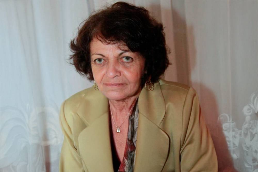 Foto de Celuta Alviano, uma mulher idosa, branca, com cabelos curtos avermelhados. Ela usa um blazer creme, uma blusa estampada e um colar.