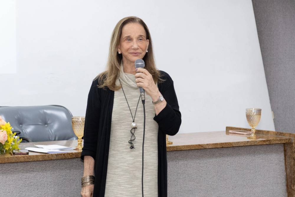 Foto de Miriam Struchiner, uma mulher idosa, branca, de cabelos loiros, que veste um vestido bege e um casaco preto.