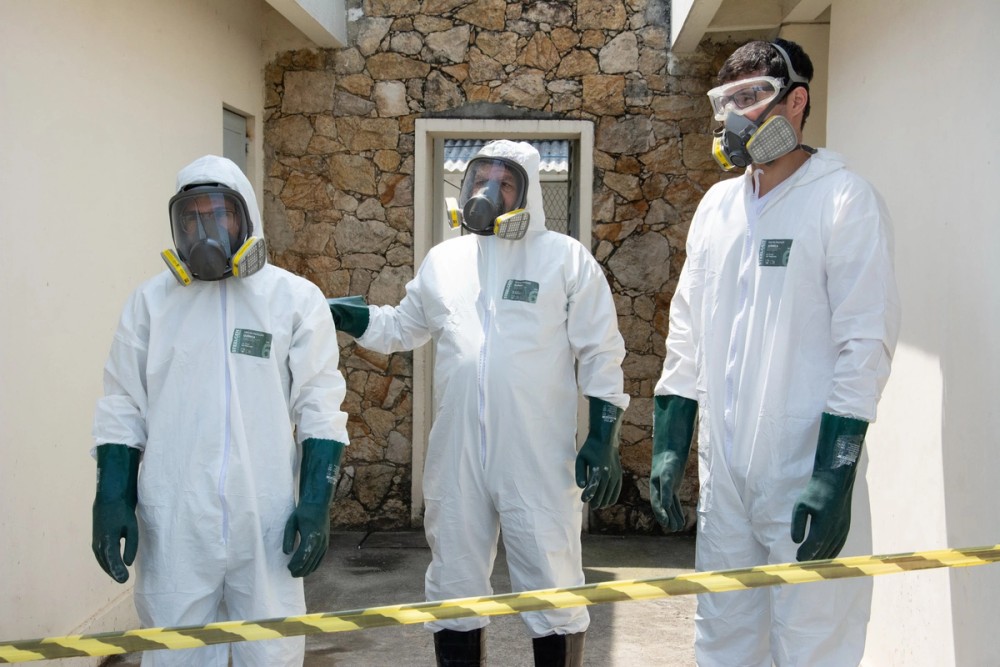 Três pessoas estão vestidas com roupas de proteção completa, conhecidas como EPIs (Equipamentos de Proteção Individual). Elas usam macacões brancos que cobrem o corpo todo, luvas grossas de borracha verde, botas pretas e máscaras de respiração com filtros amarelos e cinzas. Duas delas também têm viseiras transparentes cobrindo o rosto inteiro. Estão em um espaço estreito entre duas paredes claras, com uma parede de pedras ao fundo e uma faixa de isolamento amarela e preta na frente. A cena passa uma sensação de precaução e segurança contra contaminação.
