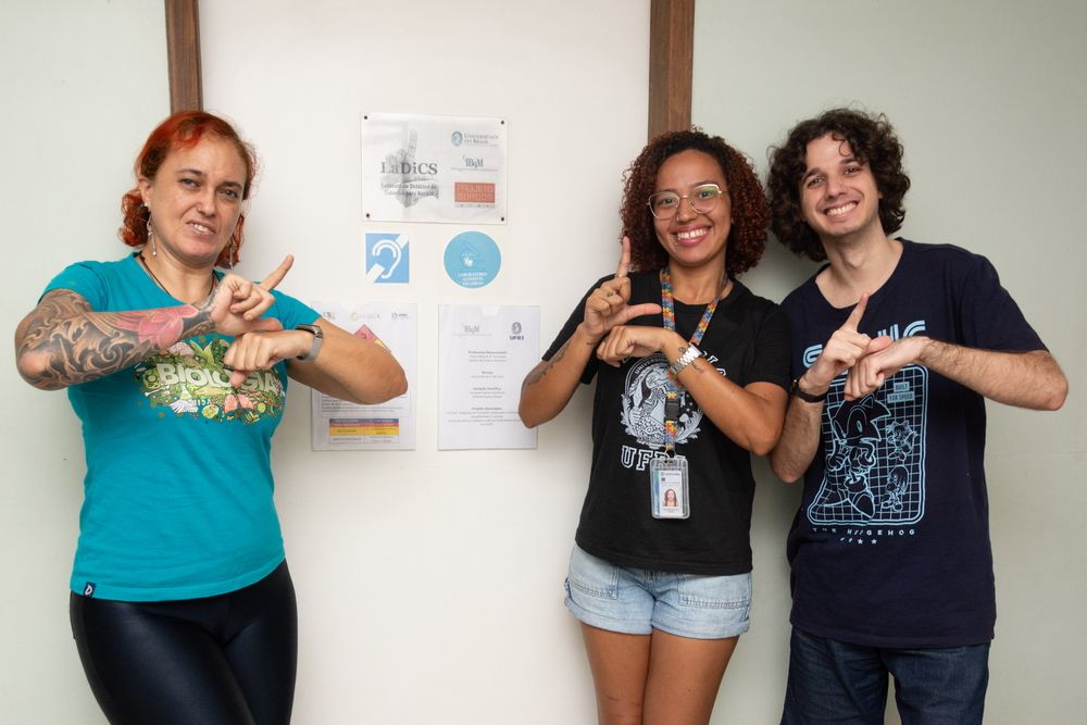 Nuccia de Cicco, Julya Marques e Michael Soares posam na porta do Ladics. Os três fazem o sinal de Libras para o nome do laboratório. 