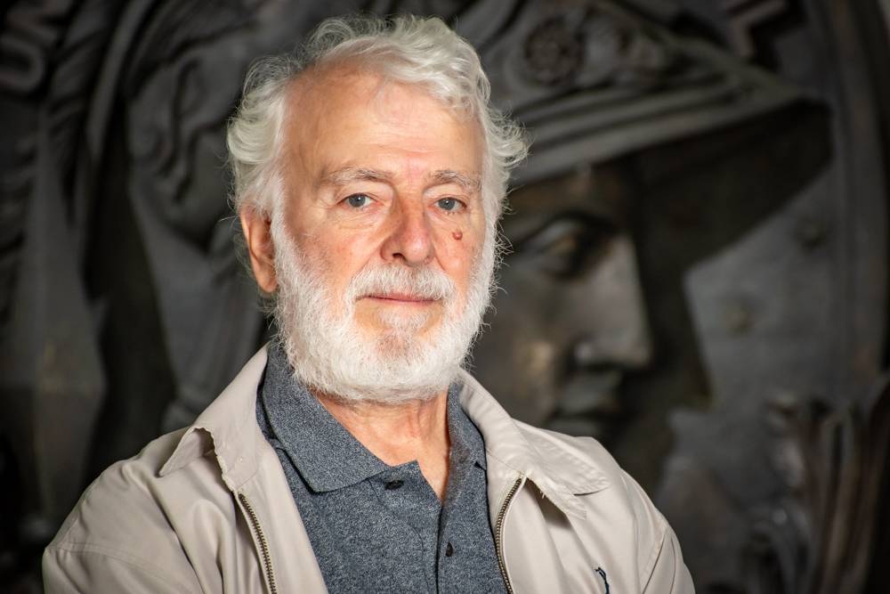 Paulo Bisch, um homem branco, idoso, com cabelos curtos e brancos. Veste uma blusa cinza e um casaco bege. No fundo aparece um pedaço da Minerva, brasão da UFRJ