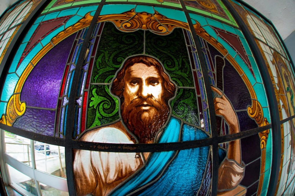 Um vitral colorido mostra a imagem de um homem de barba castanha e cabelos compridos. Ele veste uma túnica clara com um manto azul sobre o ombro. A expressão dele é séria, e ele olha diretamente para frente. O fundo é formado por desenhos geométricos e arabescos em tons de verde, roxo, azul e dourado, criando um efeito artístico e luminoso típico de vitrais.