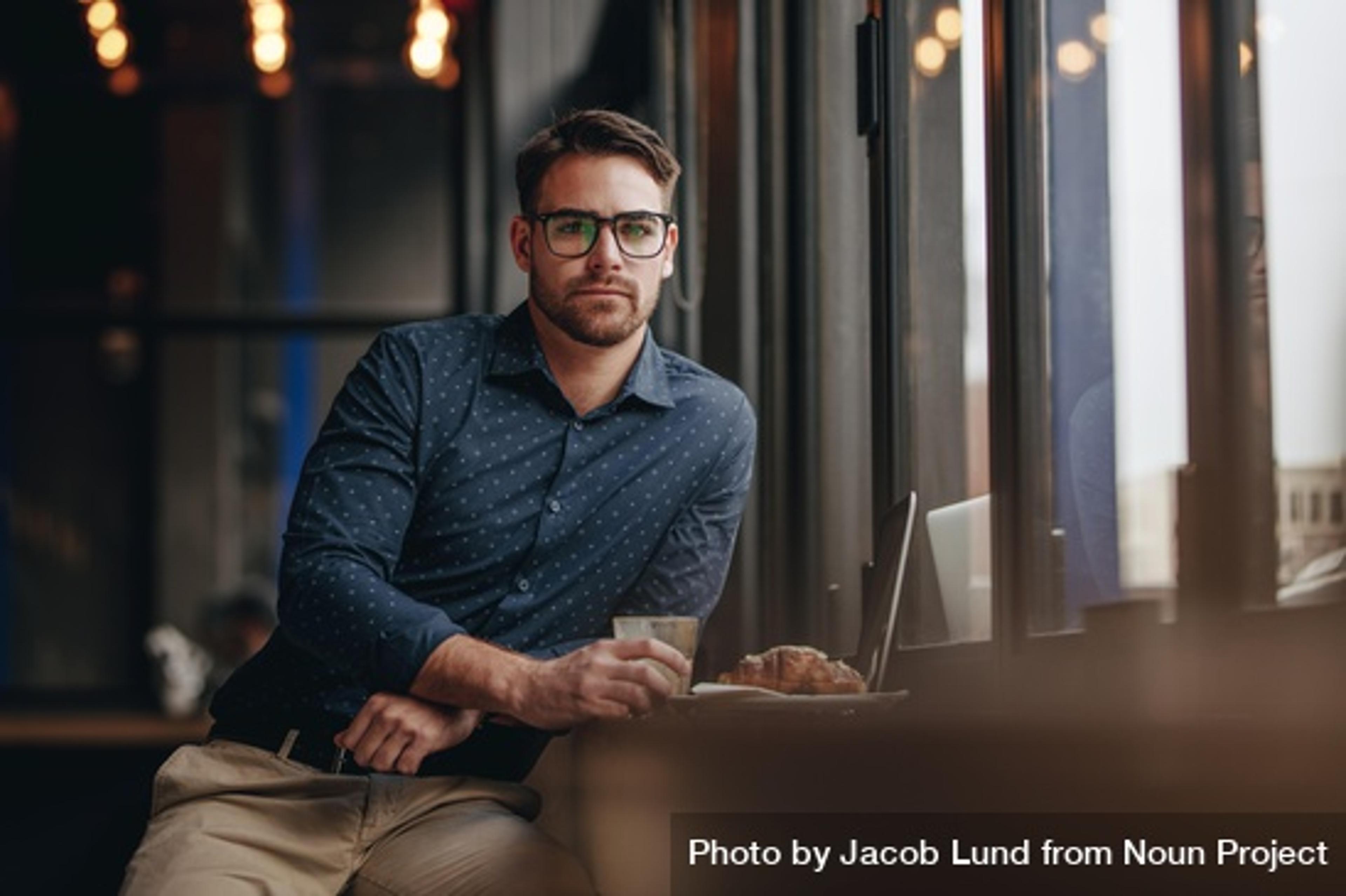 Mann med briller som sitter på kafé med laptop. Ser inn i kamera.