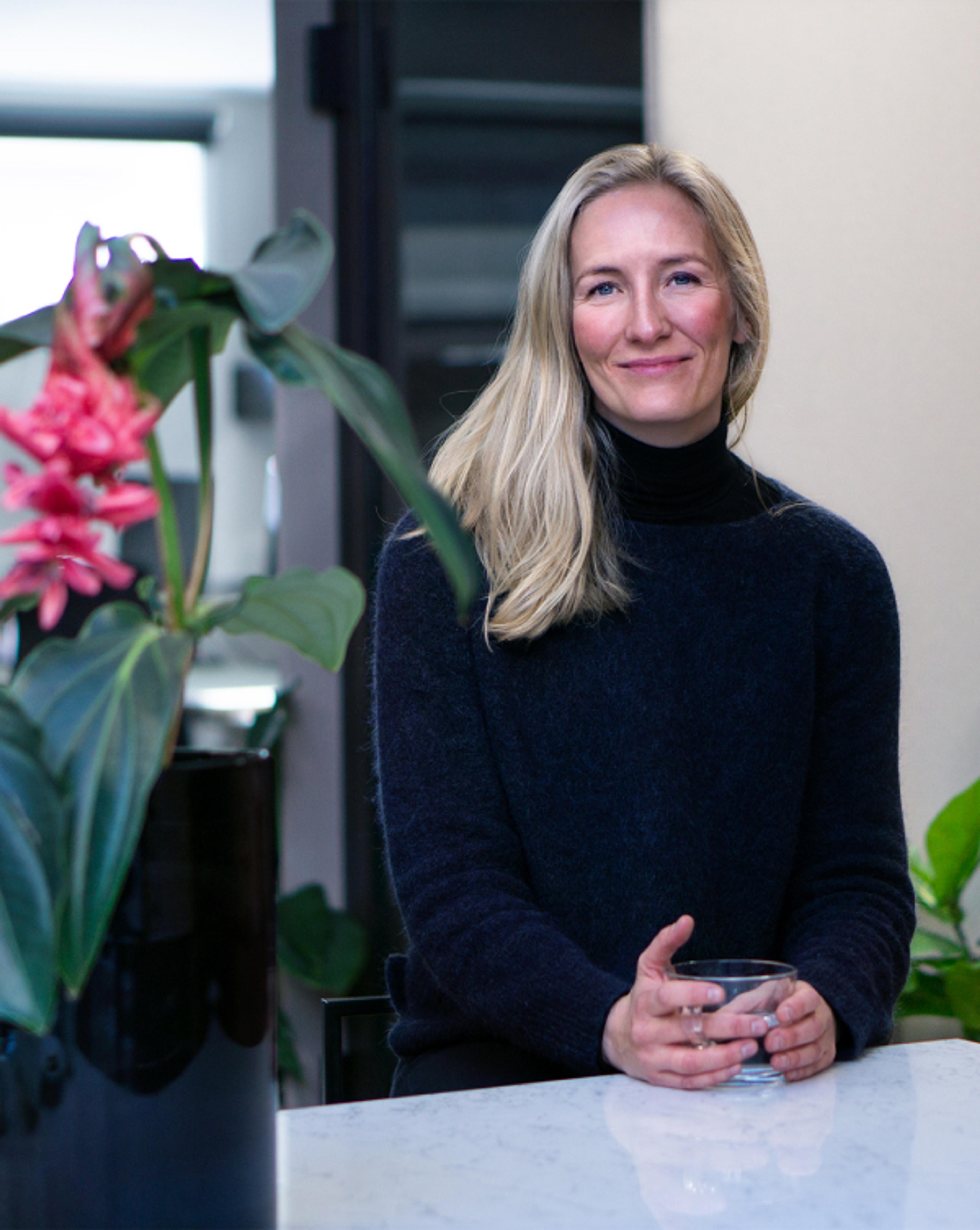 Kvinne i svart genser og med lyst hår. Sitter og smiler mens hun ser inn i kamera. Holder rundt et glass.