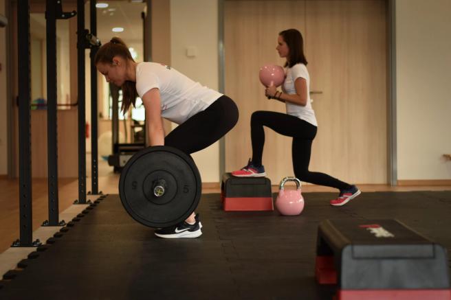 Vadeča dviguje olimpijsko palico, za njo druga vadeča z girjo stopa na stopničko.