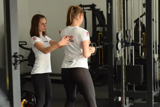 Kineziologinja popravlja držo vadeče, ki vleče ročaj na napravi s kablom.