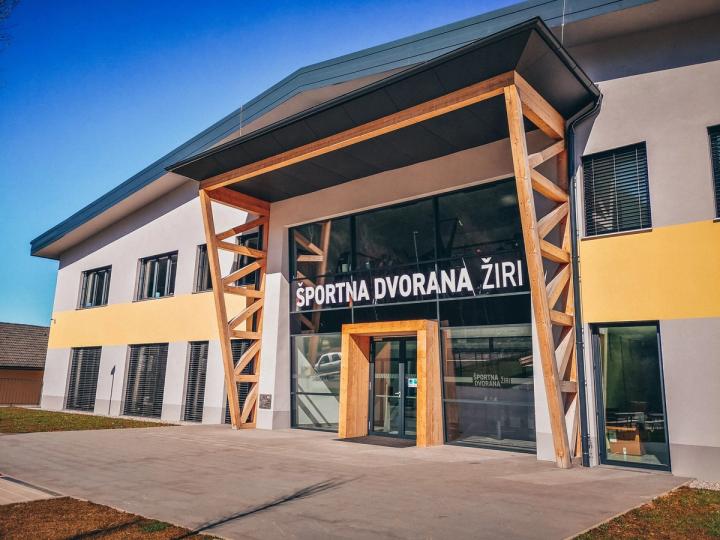 Zunanjost Športne dvorane Žiri: svetla fasada z rumenim pasom in lesenimi opornimi stebri, nad zastekljenim vhodom napis „Športna dvorana Žiri“.