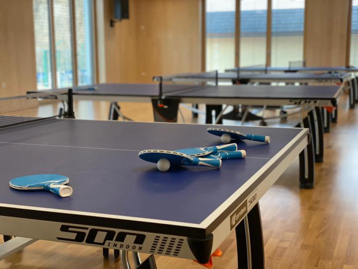 Modre mize za namizni tenis v dvorani z lesenim parketom in visokimi okni; na prvi mizi ležijo loparji in žogici.