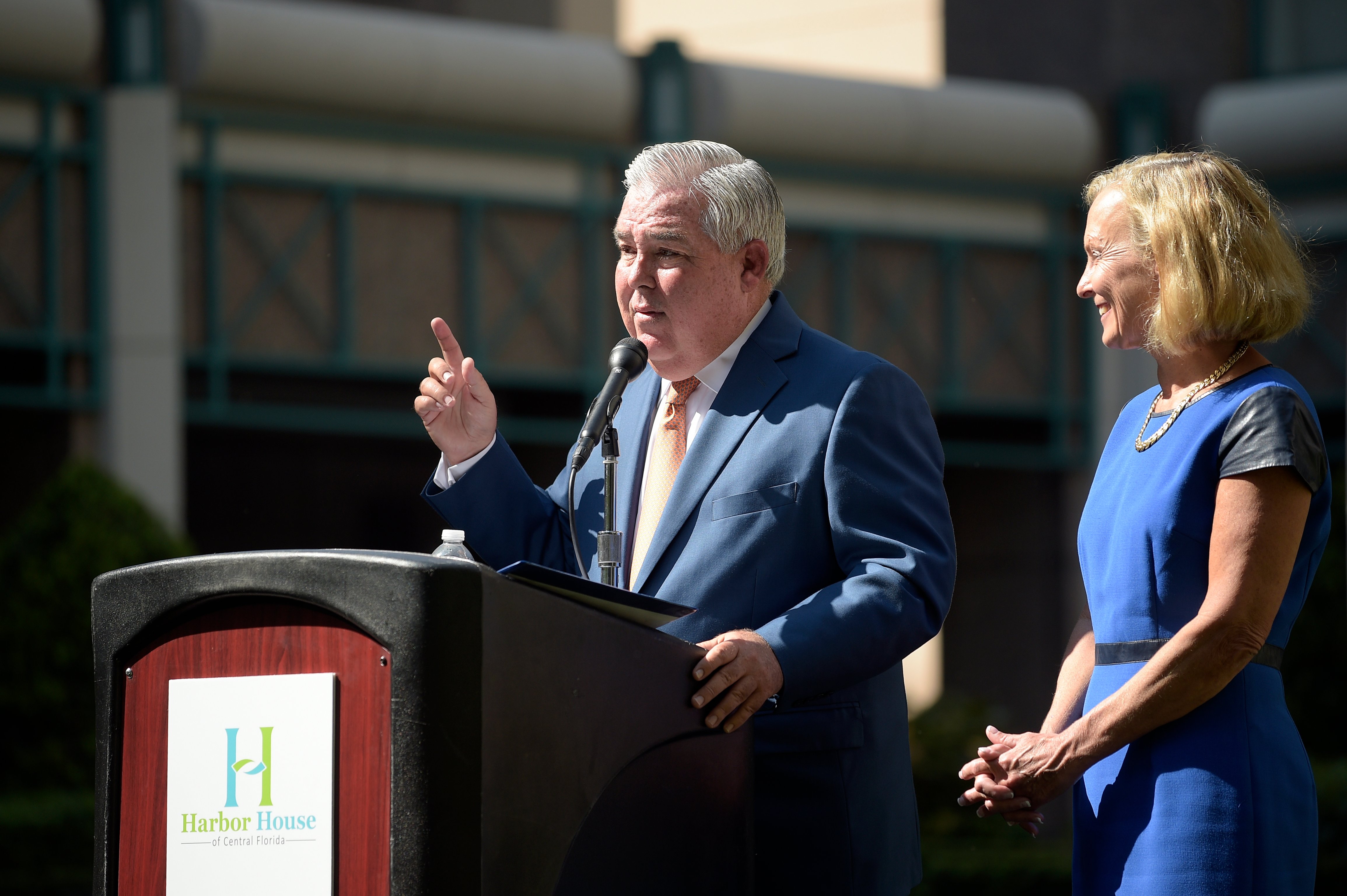 A man in a blue suit speaks at an outdoor podium while a woman in a blue dress stands beside him smiling.