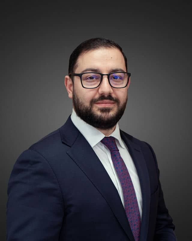 Portrait of a bearded man in glasses, a navy suit, and a purple tie against a dark gray background.