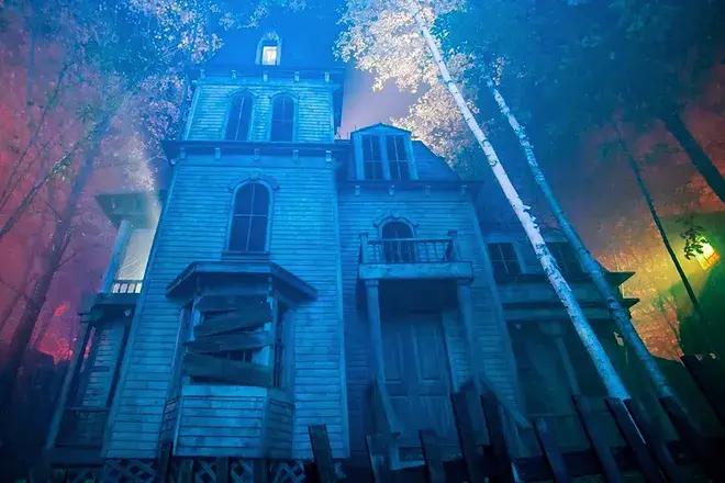 A weathered, boarded-up Victorian house looms under fog and eerie colored light among bare white trees.
