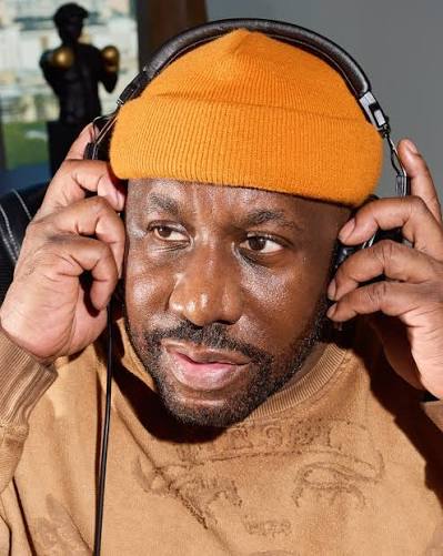 A man in an orange beanie and tan sweater holds over-ear headphones to his head with both hands.