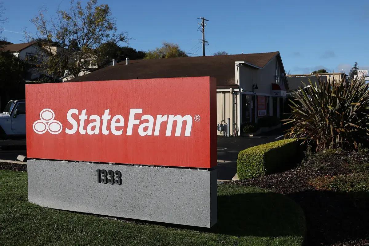 A red State Farm sign on a gray base in front of a small office building under a blue sky.
