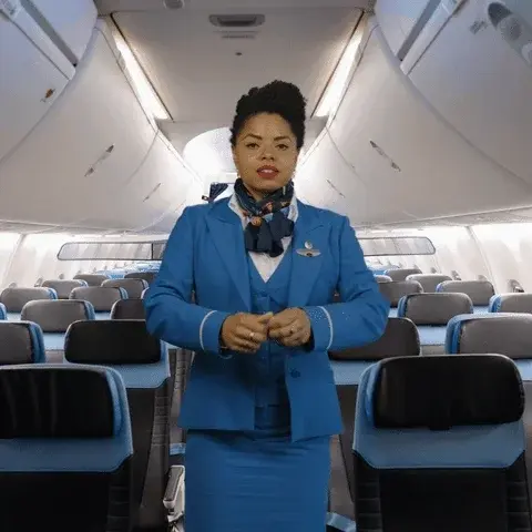 A flight attendant in a blue uniform and scarf stands in the aisle of an empty airplane cabin.