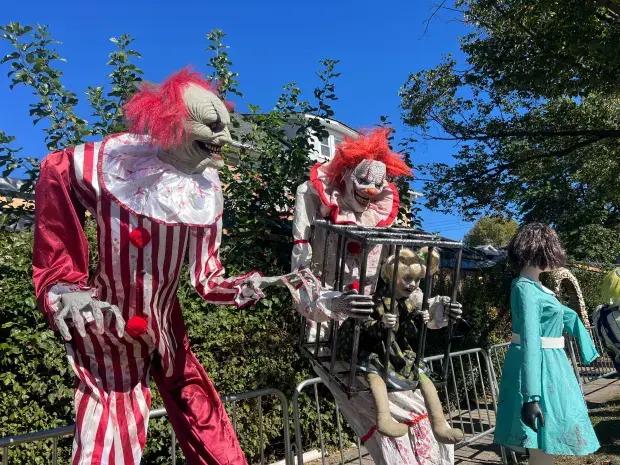 Life-size Halloween clown figures, one holding a caged doll, stand beside a bloodied mannequin on a sunny front lawn.