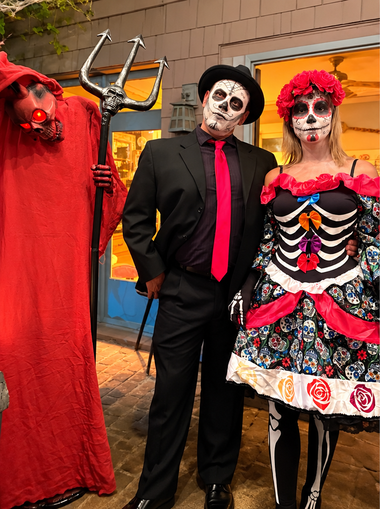 Two adults in sugar skull face paint and festive costumes pose beside a red-robed figure holding a trident.