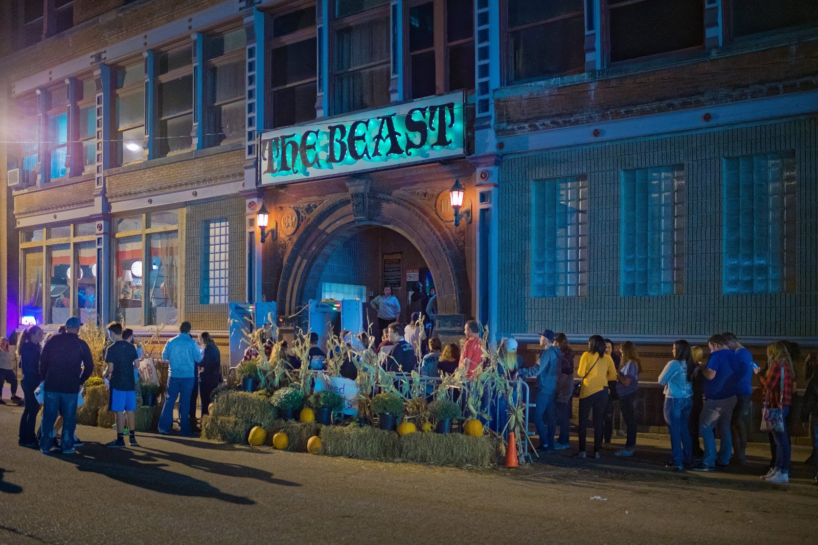 A crowd lines up at night outside a brick haunted attraction with a glowing The Beast sign and hay bale decorations.