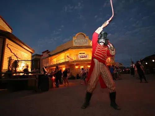 A costumed ringmaster in a skull mask raises a whip on a dusk-lit Old West street lined with a glowing hotel and saloon.