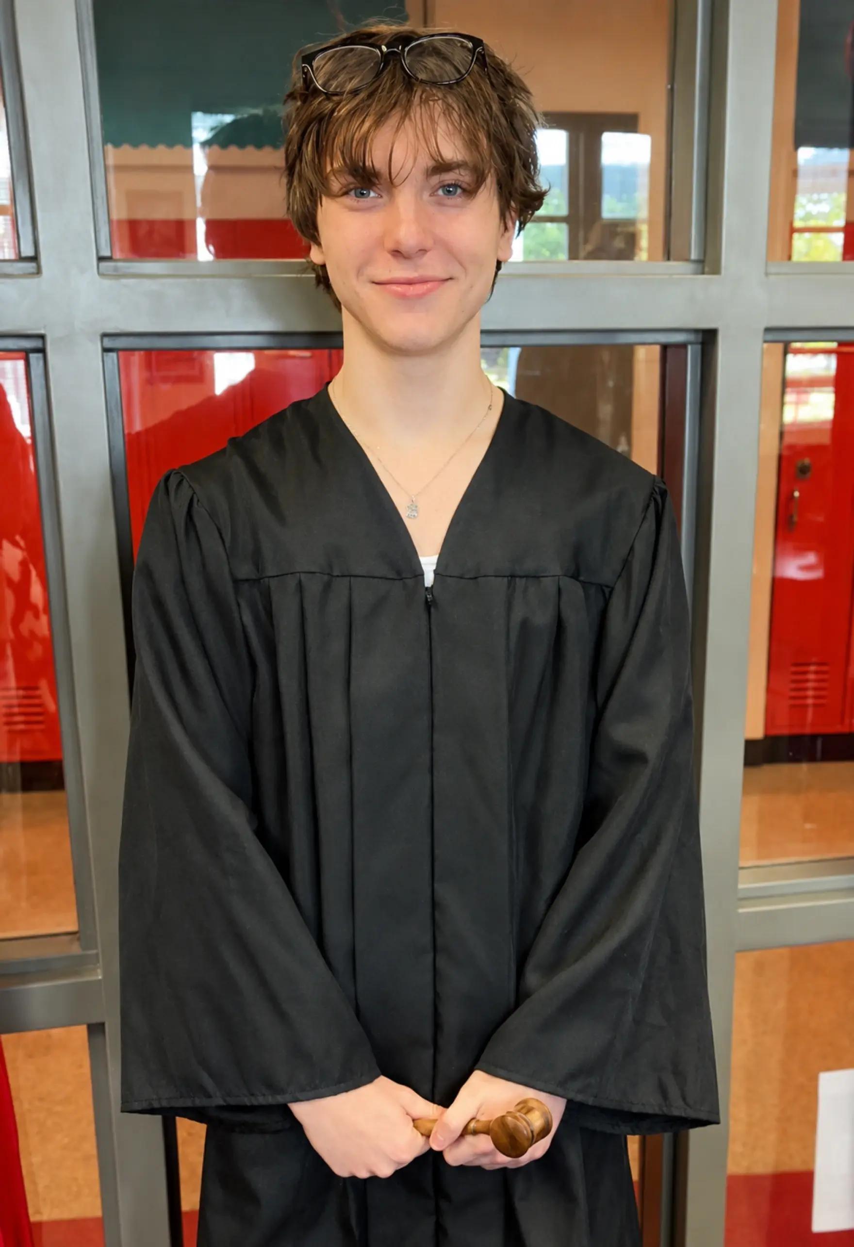 A smiling teenager in a black judge's robe holds a wooden gavel in a school hallway with red lockers.
