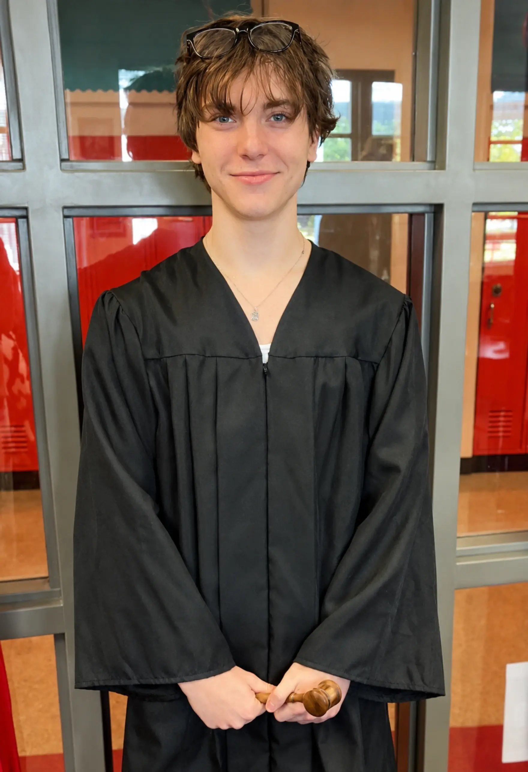 A smiling teenager in a black judge's robe holds a wooden gavel in a school hallway with red lockers.