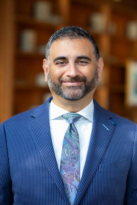 A smiling bearded man in a blue pinstripe suit and paisley tie stands in front of bookshelves.
