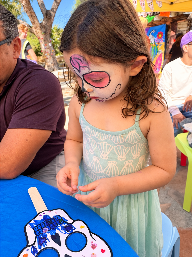 A young child with sugar skull face paint decorates a paper skull mask at a blue table outdoors.